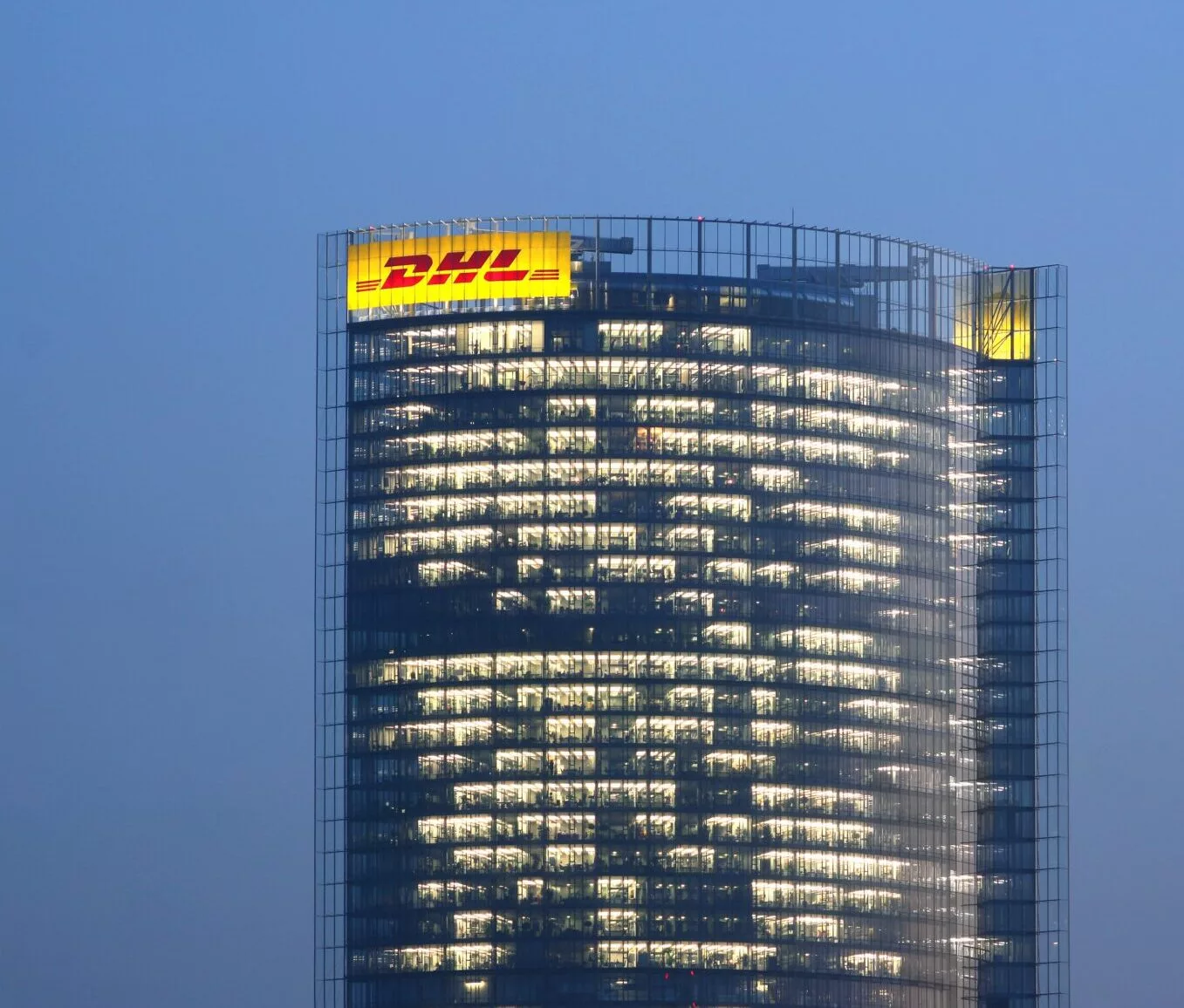 Post Tower in Bonn bei Dämmerung, beleuchtete Glasfassade mit DHL-Schriftzug an der Gebäudespitze