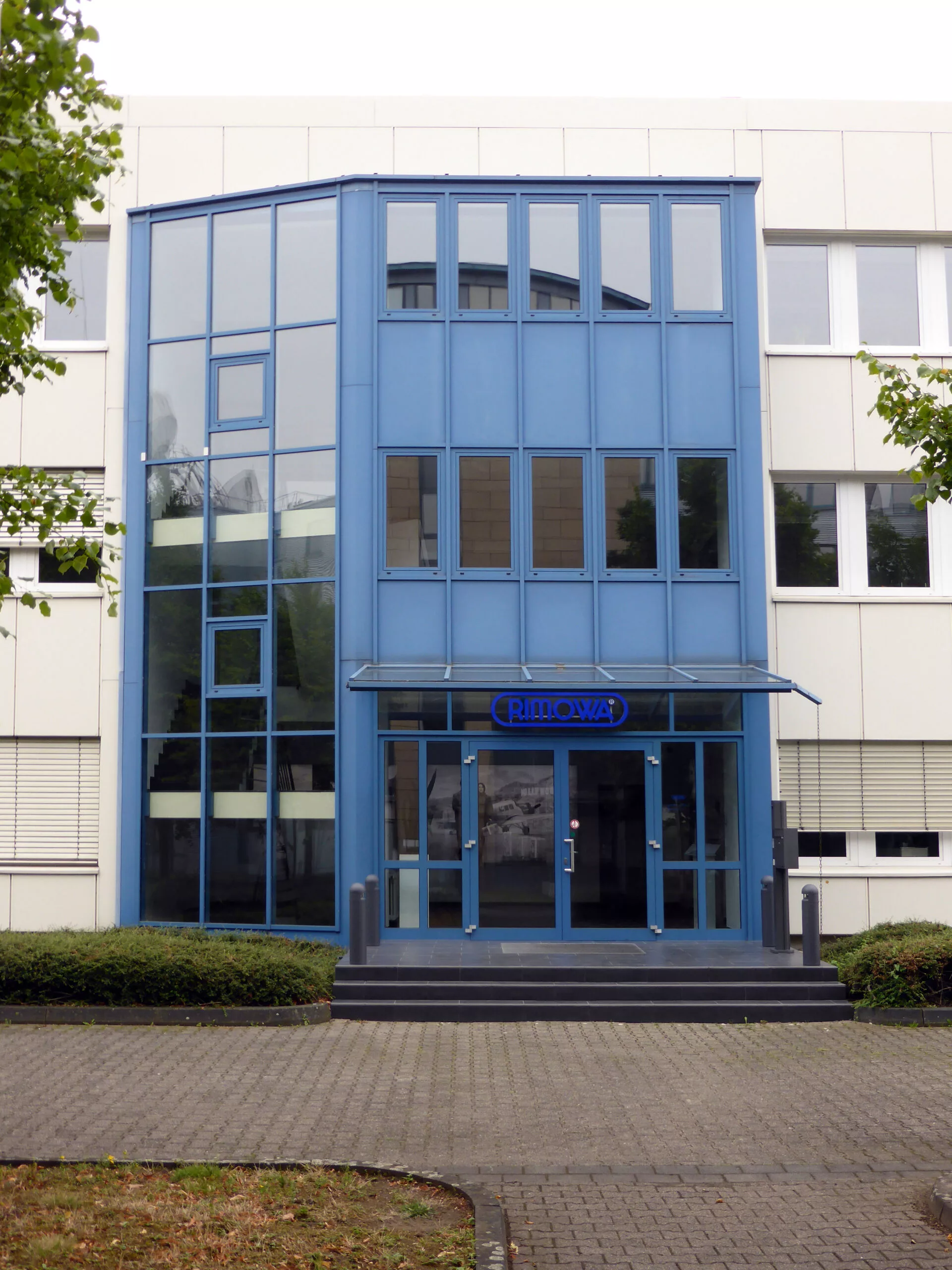 Eingangsbereich des RIMOWA Verwaltungsgebäudes mit blau gerahmter Glasfassade und Firmenschriftzug