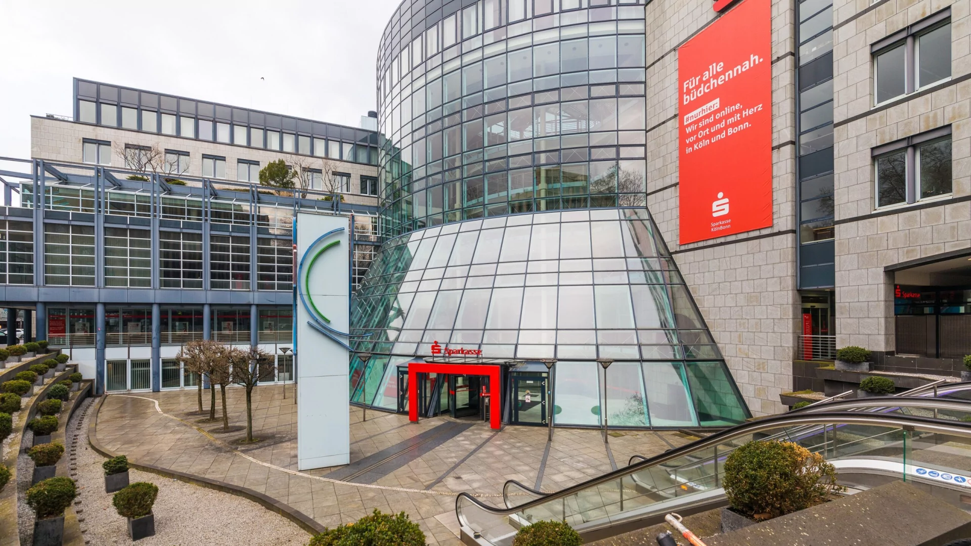 Sparkasse KölnBonn am Hahnentor in Köln mit gläsernem Eingangspavillon und rotem Banner an der Fassade.