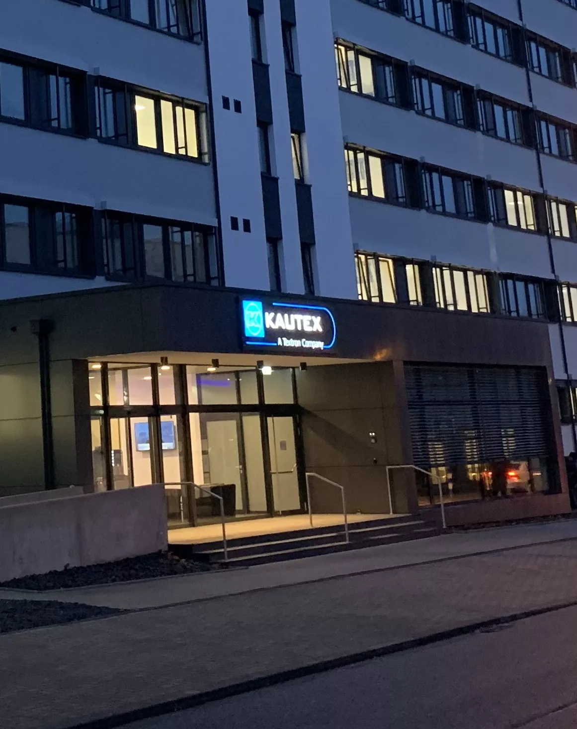 Kautex Bonn: Eingangsbereich mit Glastüren und beleuchtetem Schriftzug am Bürogebäude in der Dämmerung