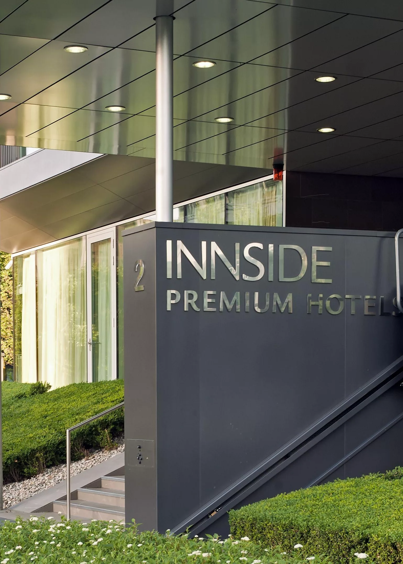 INNSIDE Premium Hotels: Eingang mit beschrifteter Stele, bodentiefer Verglasung und beschnittenen Hecken